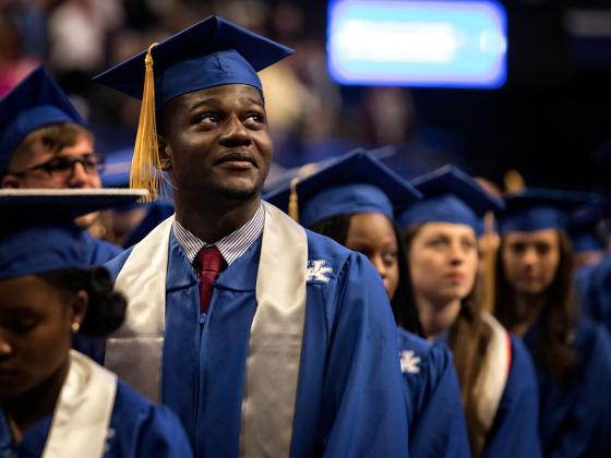 A young man in a blue graduation cap and gown with a white stole and a University of Kentucky logo stands among fellow graduates at a commencement ceremony. He gazes upward with a proud and reflective expression. Other graduates in blue caps and gowns are visible in the background, slightly out of focus, in a large indoor arena with bright lighting.