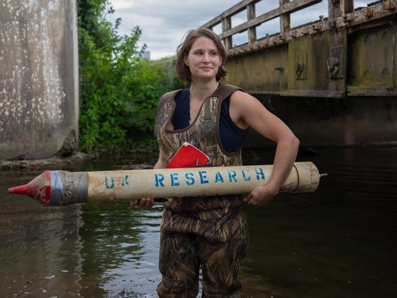Student in camo waders, standing in a river between two bridges holding a PVC rocket looking item that says "UK Research" on it.em that says "UK