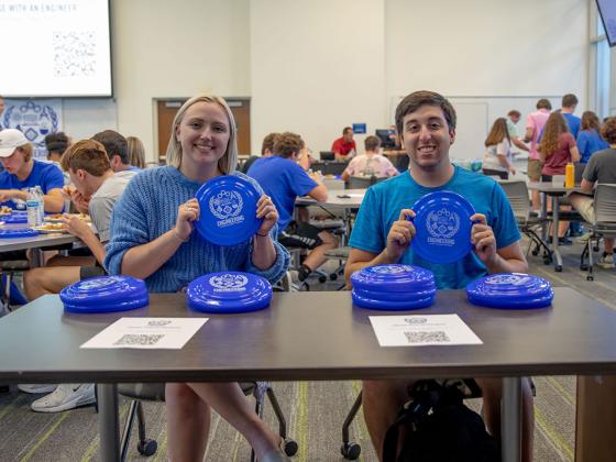 Two students holding up frisbees while sitting at a table at an event.