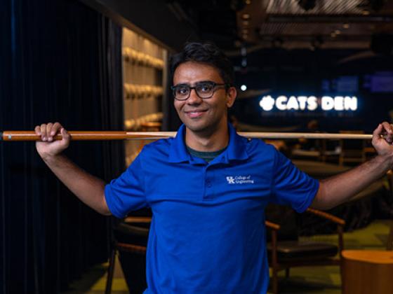 Student holding a pool queue, with a "Cats Den" sign behind him. 