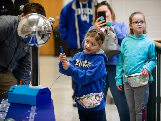 A young girl in a blue Kentucky hoodie interacts with a Van de Graaff generator, causing her hair to stand up, while people watch and take photos.