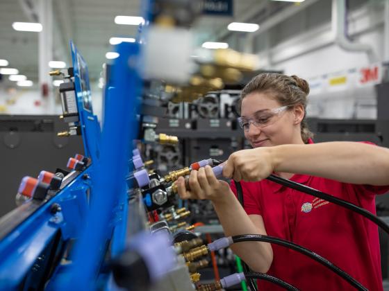A female student wearing clear safety glasses and a red polo shirt labeled "AMT" (Advanced Manufacturing Technician) works inside a bright, industrial-style training lab. She is connecting a black hose with a metallic fitting to a blue equipment panel that features multiple valves and colored connectors. The background shows rows of machinery bays, overhead lighting, and a clean, organized workshop environment focused on technical skill development.
